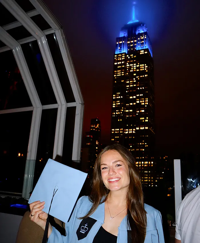 Columbia graduate Kristin Elam in graduation regalia poses with the Empire State Building from Top of the Strand Rooftop.