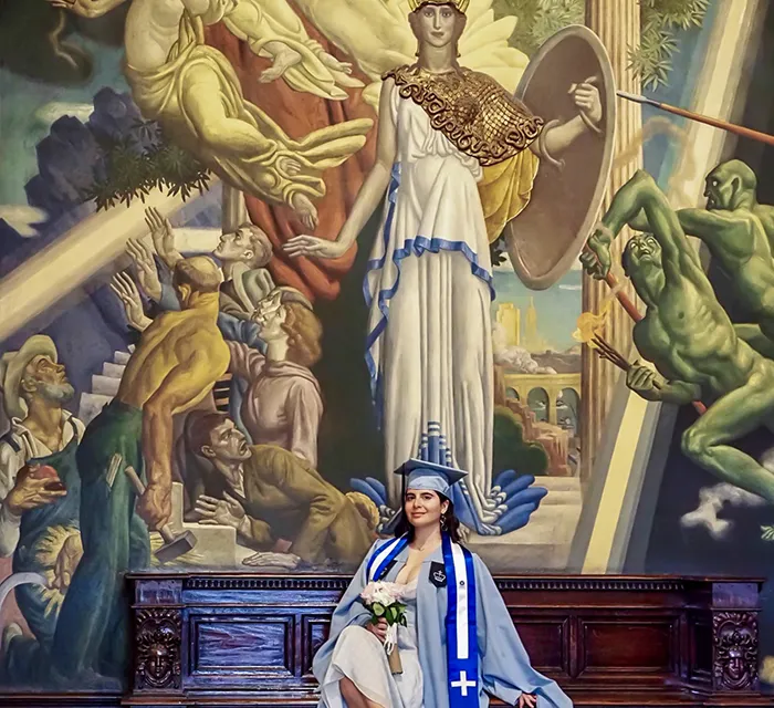 A Columbia grad poses under a mural of Athena in Butler Library. 
