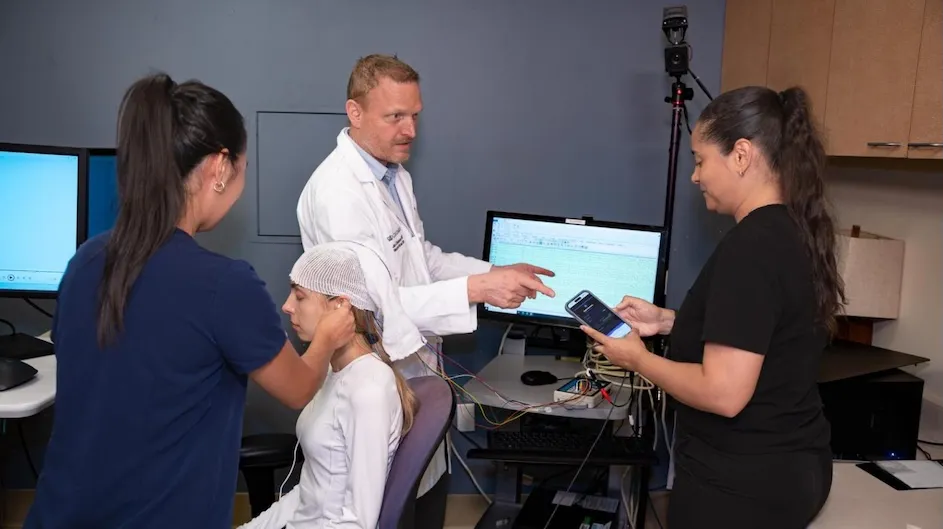 A new study shows that unresponsive ICU patients with brain injuries—when connected to a noninvasive brain-computer interface designed by Columbia researchers—were able to control the frequency of a beeping tone with their minds. Lead researcher Jan Claassen (white coat) demonstrates the interface with colleagues Sandy Song (left), Ami Camy (sitting), and Angela Velazquez (right). (Photo by Rudy Diaz/Columbia University Irving Medical Center.)