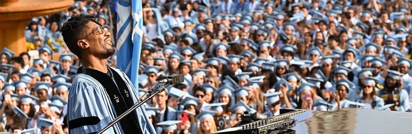Musician Jon Batiste wearing light blue Columbia regalia sings while playing at a piano in front of thousands of graduating university students in Columbia regalia on a sunny day at Columbia University. 