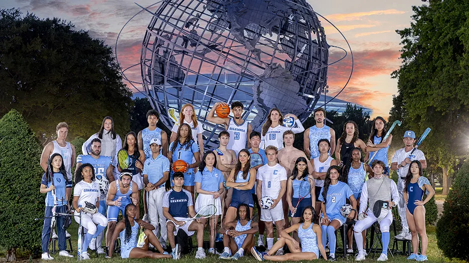 Thirty-three university student-athletes in Columbia University sports uniforms pose in front of the Unisphere in Queens, New York City, at dusk.