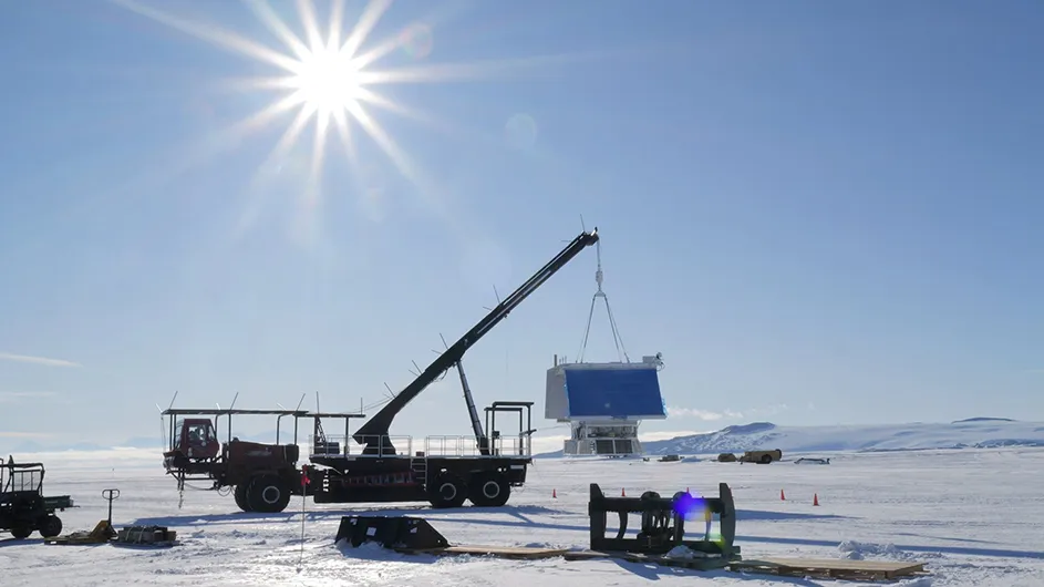 A crane lifts a large container in a snowy Antarctic landscape under a bright sun for a Columbia University research experiment.