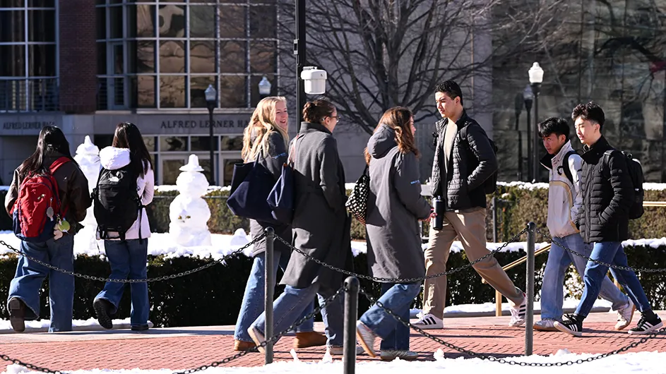 Several university students walk and converse on College Walk with snow people on South Lawn nearby at Columbia University on a sunny winter day.