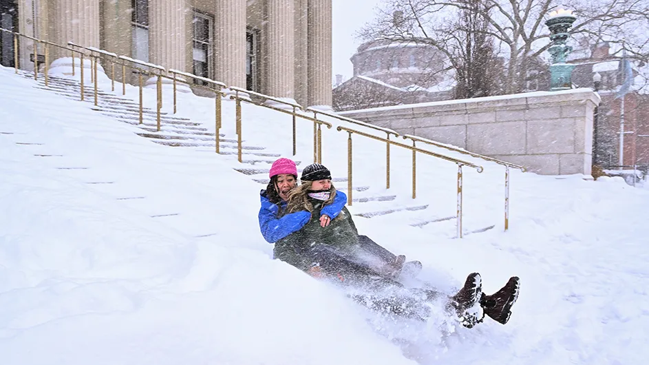 Two university students laugh joyfully while sledding down Low Library steps at Columbia University during a snowstorm.