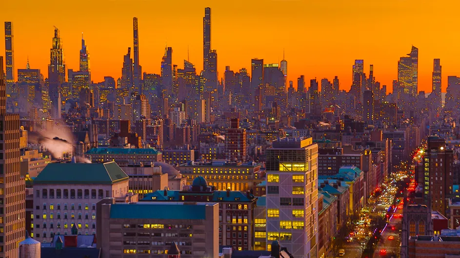 Columbia University's illuminated Morningside campus with New York City's skyscraper skyline in the distance against an orange-colored sky at dusk.