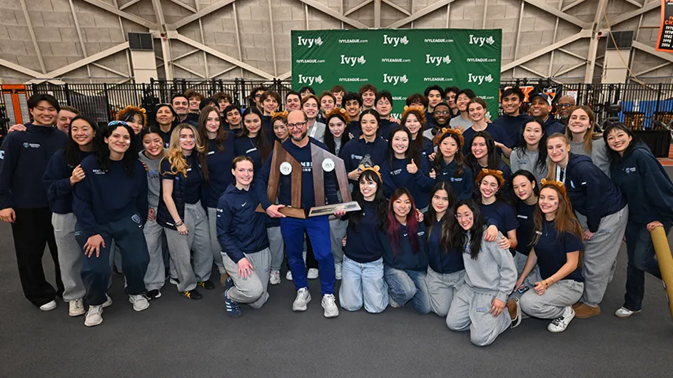 Several Columbia University student-athletes and coaches in athletic uniforms smile and pose while celebrating with a trophy at the Ivy League Fencing Championships.