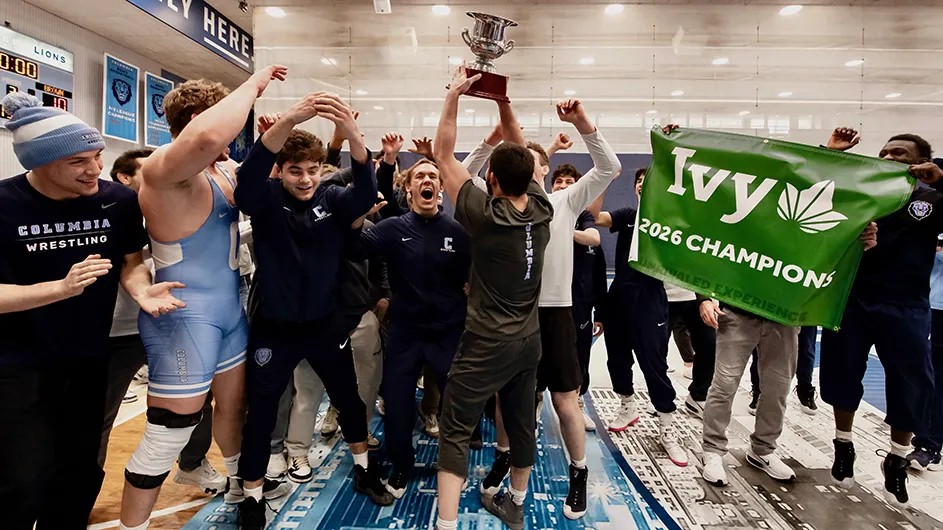 Several Columbia University student-athletes and coaches in athletic uniforms cheer and smile while celebrating with a trophy for the Ivy League Wrestling Championships.