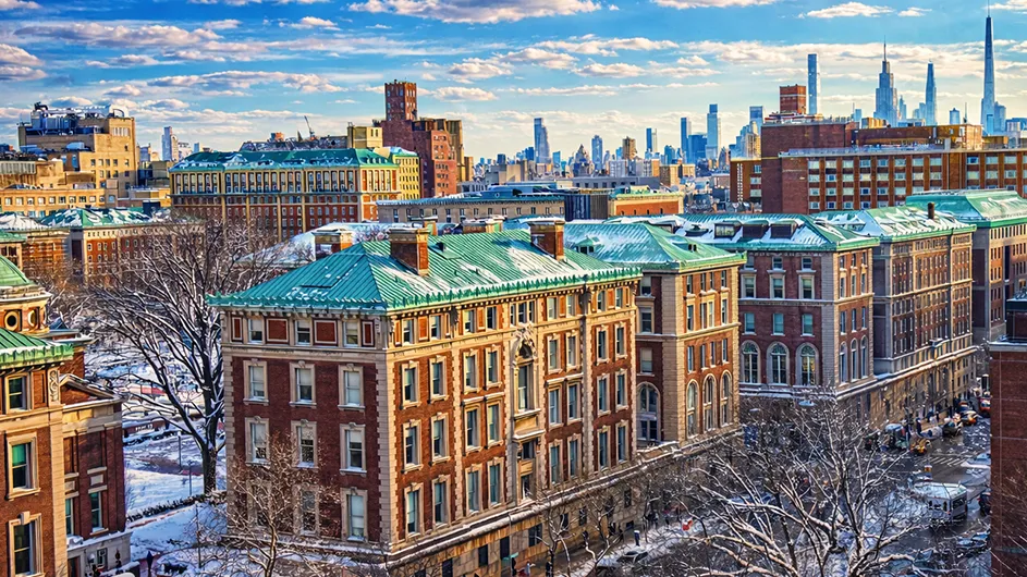 Skyline view of Columbia University Morningside campus with red-brick buildings and green rooftops as well as snow covering the grounds and Manhattan's skyscraper skyline in the background under a partly cloudy sky.