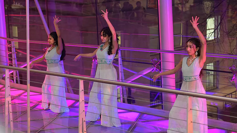 Three university students wearing long flowing white dresses dance in unison with raised graceful hands on illuminated pink and purple ramps at Lerner Hall for Glass House Rocks at Columbia University at night.