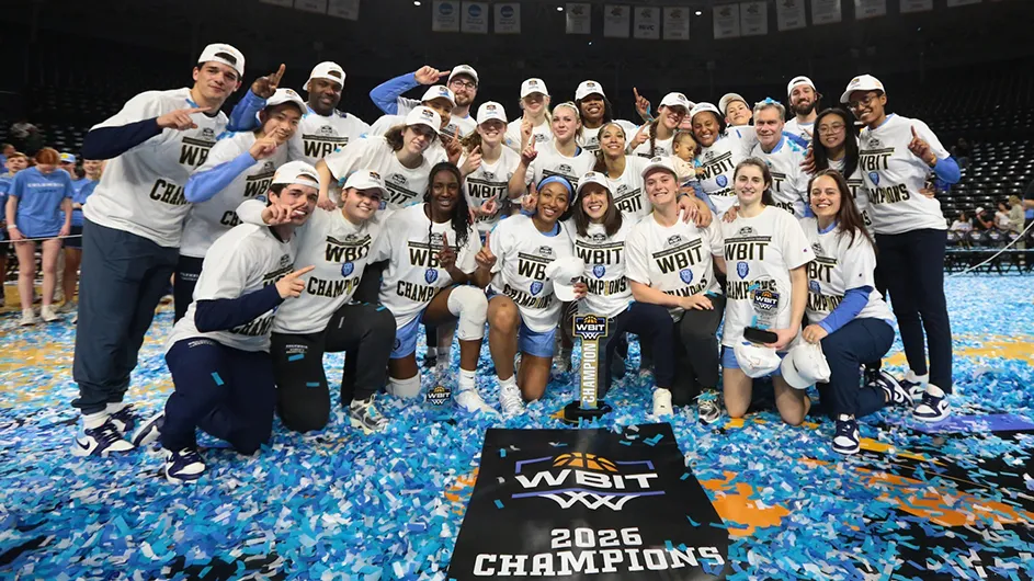 Several Columbia University students and coaches in basketball uniforms and WBIT Champions t-shirts smile as they hold up their index fingers behind a trophy and signage for 2026 WBIT Champions on a basketball court scattered with light blue and white confetti.