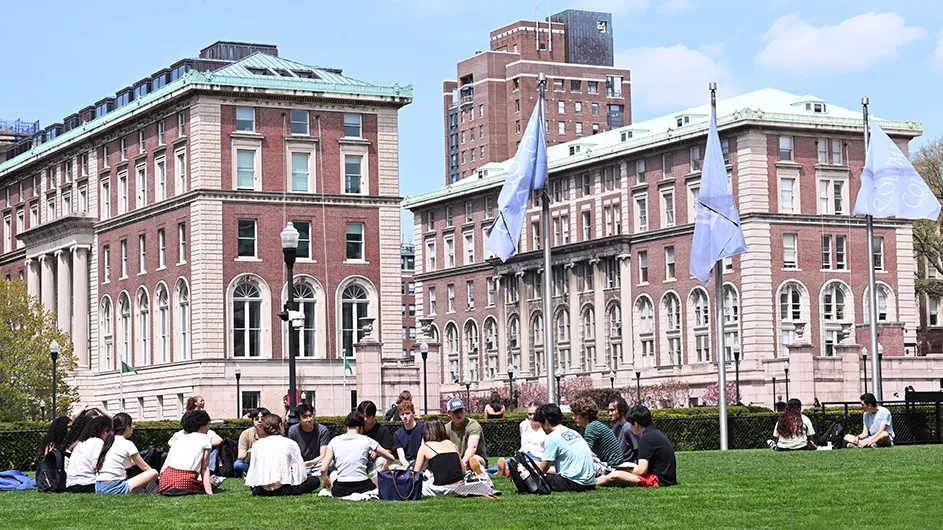 A professor and several university students sit on grassy South Lawn and engage in a class circle setting with Pulitzer Hall and Dodge Hall in the background on a sunny spring day at Columbia University.