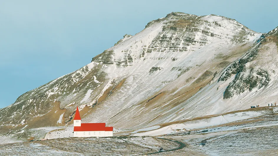 A white church with red roof and tower against snowy mountains in Iceland on a sunny day.