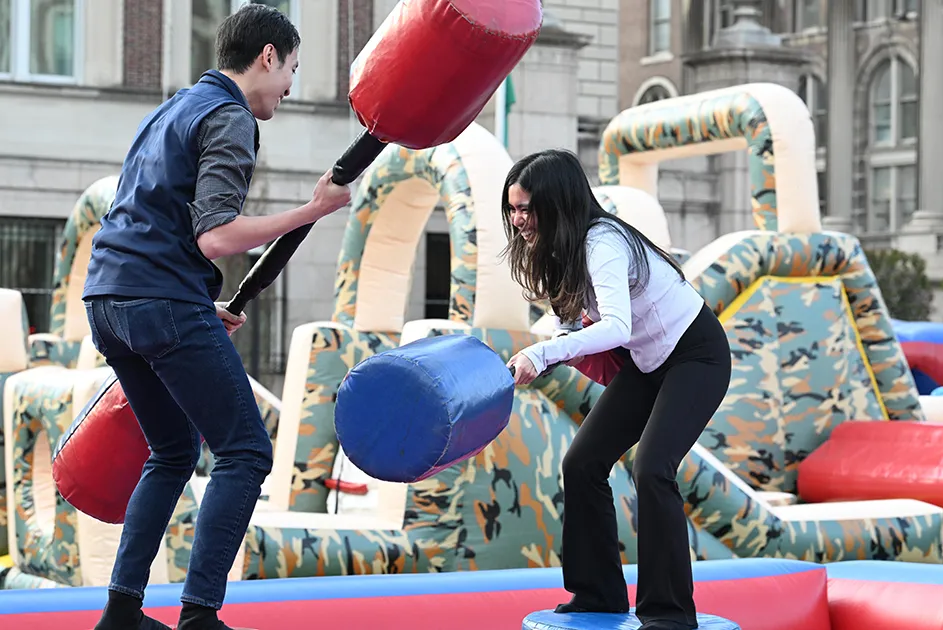 Two university students laugh and play with inflatable jousting hammers to knock each other off raised pedestals at Night Carnival event at Columbia University.
