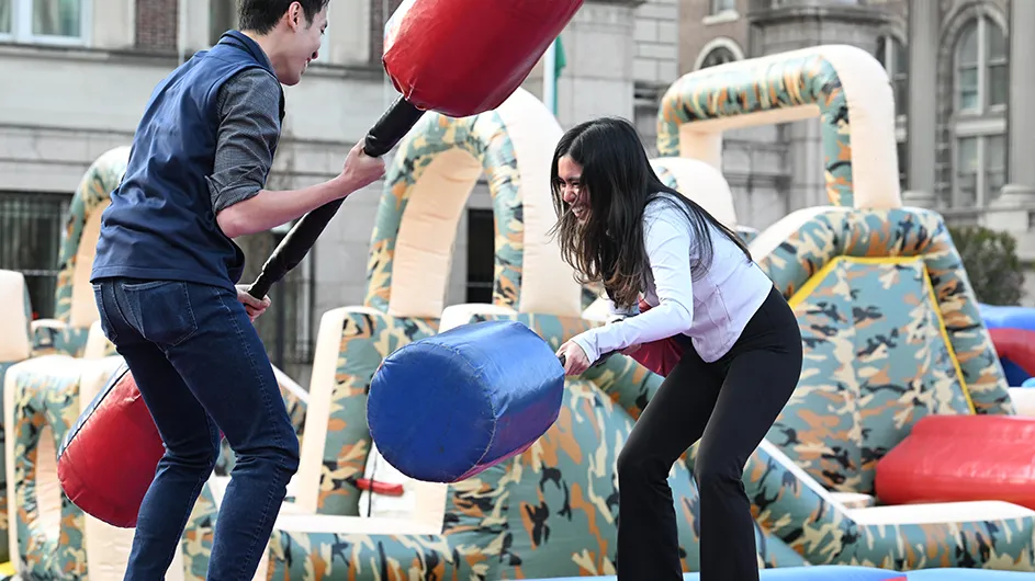 Two university students laugh and play with inflatable jousting hammers to knock each other off raised pedestals at Night Carnival event at Columbia University.