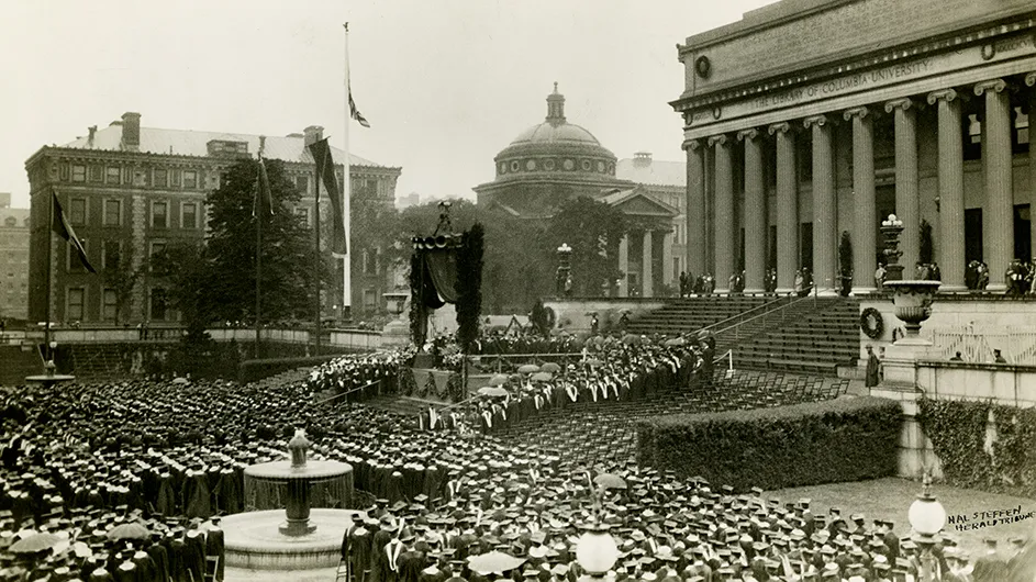 Hundreds of graduating university students and faculty in caps and gowns are seated at Columbia University’s 1926 graduation Commencement on Low Plaza with Low Library, Earl Hall, and Lewisohn Hall in the background at Columbia’s Morningside campus. 