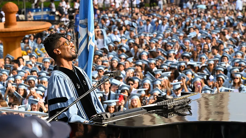 Musician Jon Batiste wearing light blue Columbia regalia sings while playing at a piano in front of thousands of graduating university students in Columbia regalia on a sunny day at Columbia University. 