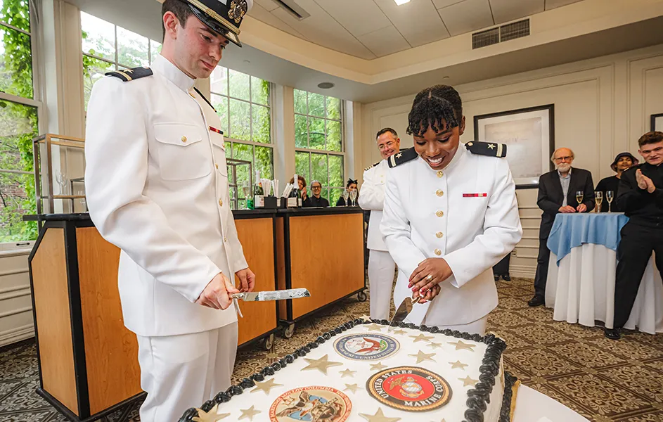 Two Columbia graduating students (Dana Clarke and Paul Randall) dressed in white naval uniforms cut a ceremonial cake with knives at their NROTC commissioning ceremony at Columbia University Faculty House.