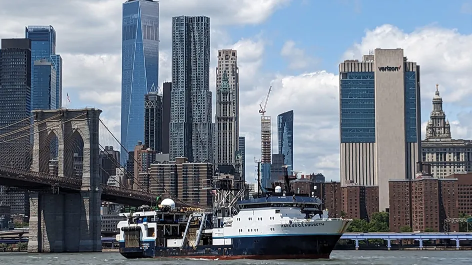Columbia University’s Research Vessel (R/V) Marcus G. Langseth in the East River with the Brooklyn Bridge, World Trade Center, Manhattan Municipal Building, and the downtown New York City skyline in the background on a partly cloudy day. 