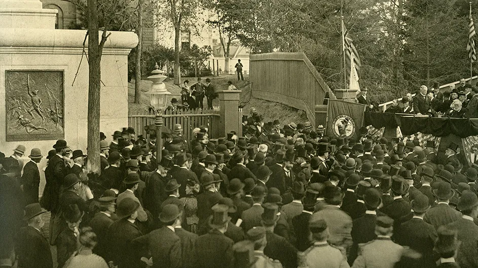 A ceremony in 1897 with dozens of spectators in formal wear in front of a stage adorned with American flags as a speaker addresses Columbia President Butler with the dedication of a bronze plaque commemorating the 1776 Battle of Harlem Heights on a building wall at Columbia University.