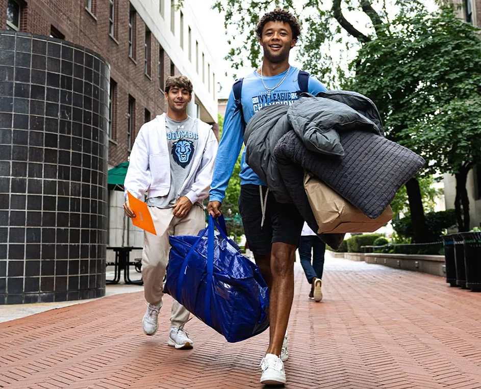 Two university students wearing Columbia University Lions shirts smile while walking with bags during move-in.