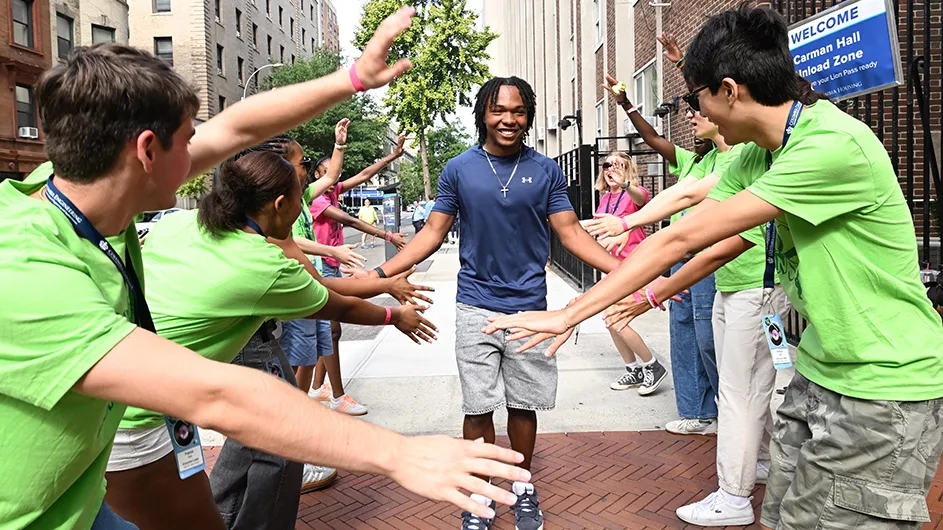 A smiling university student walks through a welcome tunnel of several cheering university students giving high-fives and low-fives as welcome gestures during Columbia University move-in.