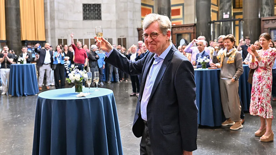 Columbia Professor Michael Woodford, in a dark suit, smiles and raises a toast of champagne with a crowd raising their glasses and clapping in the background for Woodford's Nobel Prize celebration in Low Library Rotunda at Columbia University.