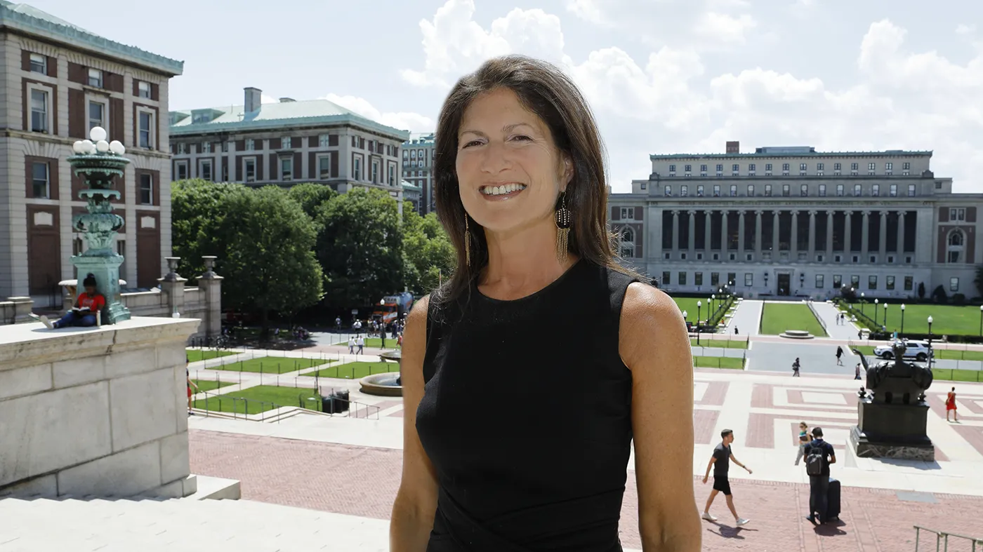 A woman in a sleeveless black dress stands in front of buildings.