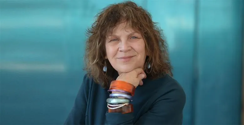 Image of Carol Becker wearing a dark blazer hand underneath chin while wearing multi colored bracelets