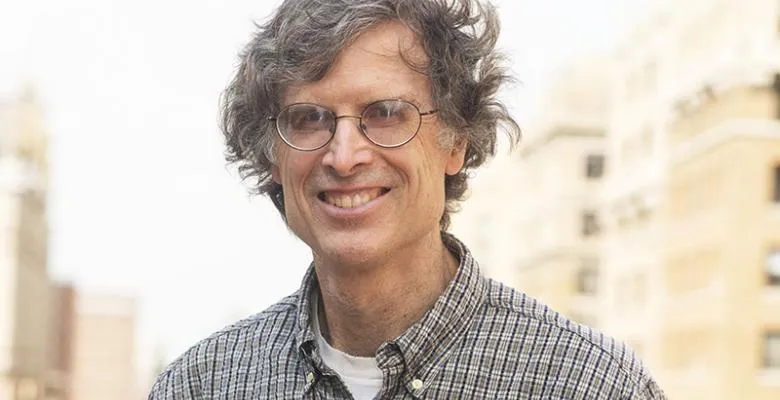 Dan O'Flaherty smiles with dark rimmed eyeglasses and a dark brown and tan plaid shirt with a white t-shirt peeping out the collar area