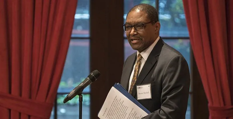 Dr. Mitchell in dark suit folder under his arm standing in front of opened burgundy drapes at a podium