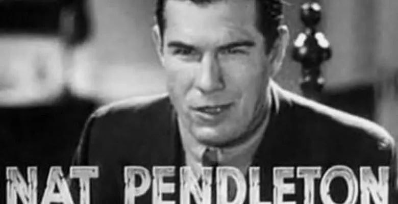 black and white image of Nat Pendleton looking directly into camera with a smirk on face dressed in a dark suit and light colored button down shirt underneath
