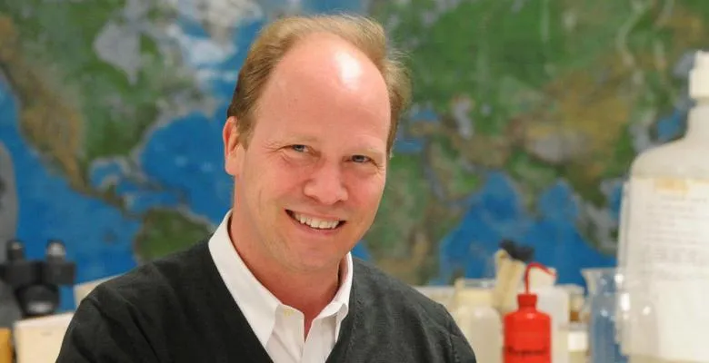 A man smiles into the camera, while standing in front of a wall map of the Earth.