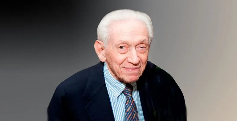 A man with white, close-cropped hair smiles into the camera against a dark gray background.