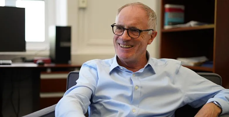 Relaxed in a dress shirt, a man sits in an armchair, smiling as he speaks to someone off-camera