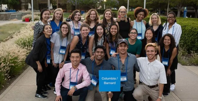 A group of roughly two dozen people poses on a sidewalk with a placard reading "Columbia/Barnard"