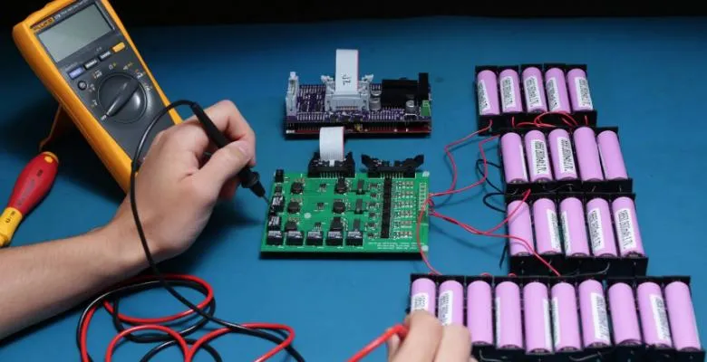 A pair of hands holds an ammeter, with one lead touching a battery pack and the other touching a motherboard