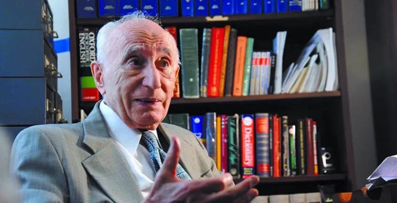 Wearing a gray suit and tie, a man sits in front of shelves lined with books, gesturing with his hands