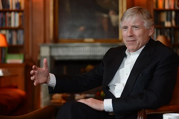 A man wearing a suit but no tie sits in an armchair in a room lined with bookshelves, gesturing with one hand as he speaks