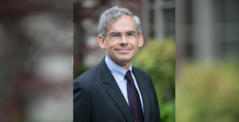 Outside on a sunny day, a man, wearing glasses and a dark suit and tie, smiles into the camera.