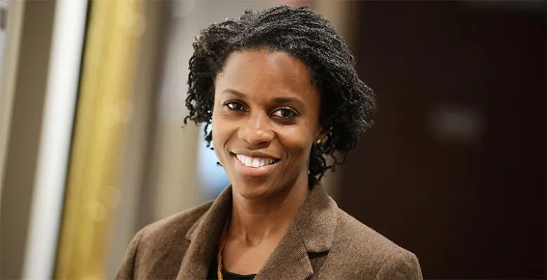 Wearing a brown, wool blazer, a woman, with her hair pulled back into a loose ponytail, smiles into the camera
