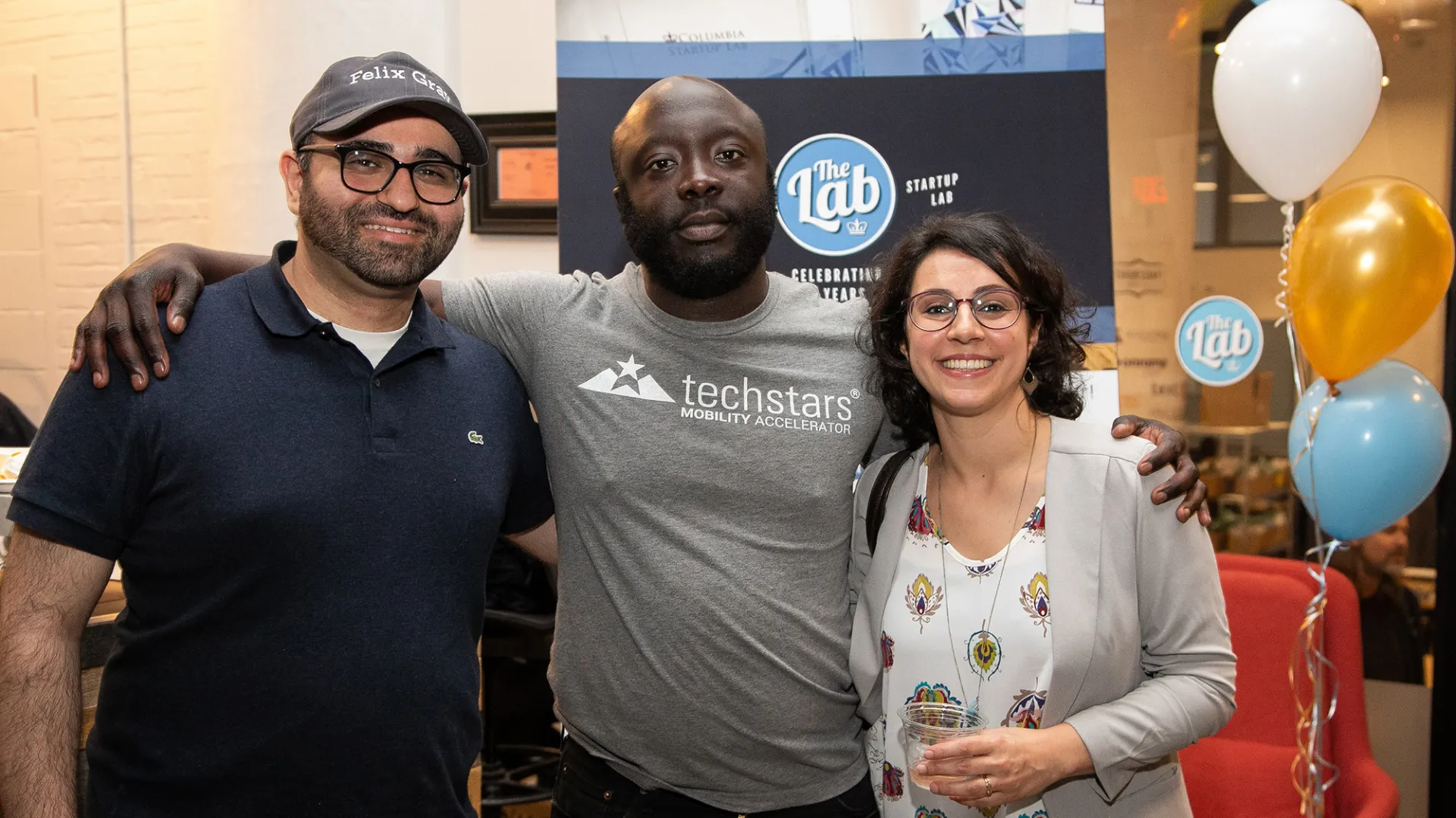 Three people with arms around each other with festive balloons in the background: Man with glasses and beard in blue collared shirt and gray baseball hat; bearded man with gray T-shirt; and woman with glasses and white floral design shirt and light gray blazer. 