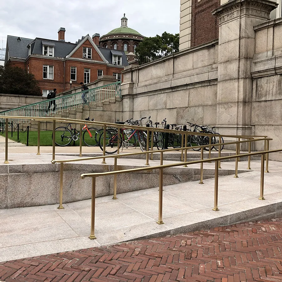 A masonry ramp with bronze rails zig zags towards a second level leading up to a staircase. 