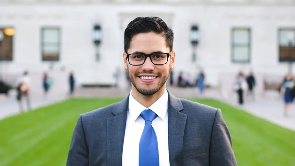 Brian Ruiz in a navy blue suit and tie, standing on a green lawn in front of Butler Library.
