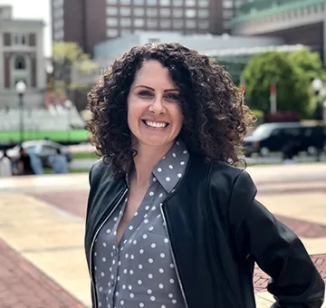 Tara Heidger in a polka dot blouse and black blazer, standing on College Walk.