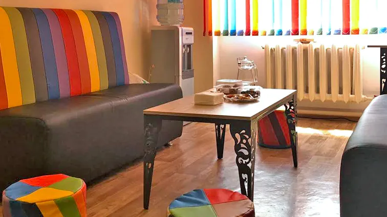 A rainbow-themed therapy room with a sofa, tufts, coffee table, a water cooler, and rainbow curtains.