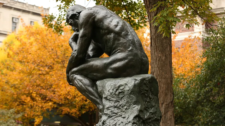 A statue of a thinking man in front of trees with fall foliage.