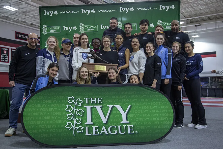 22 fencing athletes and coaches pose with a championship trophy against a green Ivy League athletics backdrop in a fencing room.