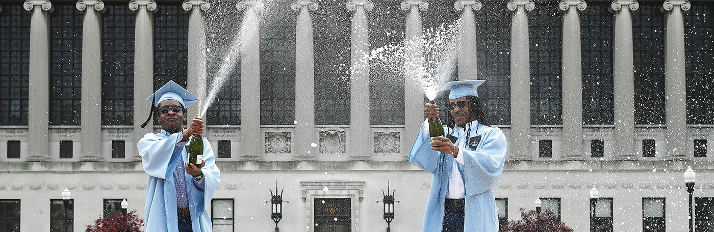 Two Columbia graduates in blue caps and gowns pop champagne in front of Butler Library.