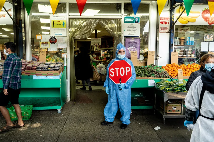 A person in full-body Personal Protective Equipment, mask, and face shield stands with a stop sign at an entrance to a New York City bodega.