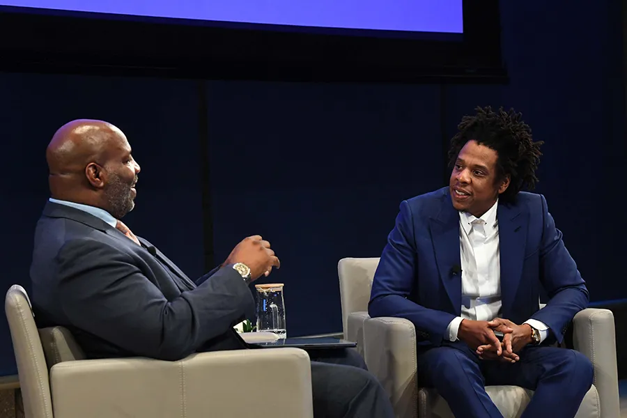 Professor Jelani Cobb and Shawn "JAY-Z" Carter sit in tan chairs in front of a black backdrop in an auditorium at The Forum.